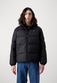Sort pufferjakke med lynlås fortil, med en quiltet design, elastiske manchetter og et lille logo på venstre bryst. Bæret over en brun sweater.