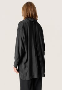 Mujer con cabello hasta los hombros que lleva una blusa negra de manga larga y holgada con un detalle de lazo en la parte posterior, combinada con pantalones negros.