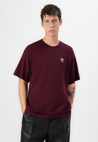 Jauns vīrietis ar īsiem tumšiem matiem, tērpies bordo Adidas T-krekliņā un melnās biksēs, stāv ar rokām kabatās uz balta fona.