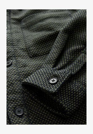 Textured dark green fabric with small checkered pattern, featuring a folded sleeve and black buttons. Notable stitching detail.