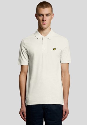 Jonge man die een effen gebroken witte polo draagt met een klein geel vogel-logo op de linkerborst en donkere broek, staand tegen een grijze achtergrond.