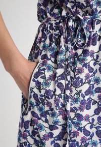 Vestido floral con fondo blanco y patrones en púrpura, verde azulado y rosa. Incluye cintura ajustada y bolsillos laterales con cremallera.