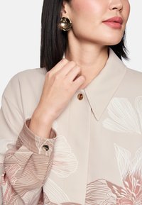 Long-sleeve blouse in soft beige with floral patterns. Collared neckline, button closure, and metal accent on cuff. Round gold earrings.