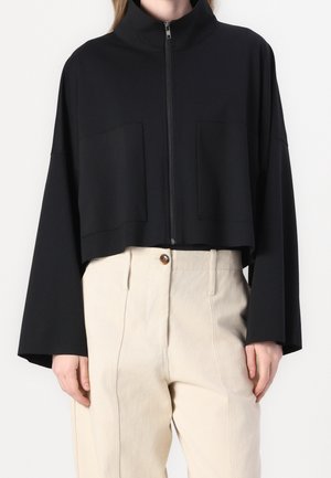 Person, die eine schwarze, kurze Jacke mit Reißverschluss und langen Ärmeln sowie beige, hoch taillierte Hosen mit Knopfverschluss trägt.