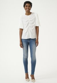 Blusa bianca con maniche a palloncino e arricciature sul davanti, abbinata a jeans skinny blu e scarpe beige con dettaglio a nodo. Sfondo semplice.