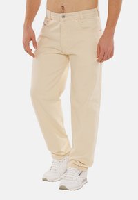 Beige Jeans aus Denim mit geradem Bein, ausgestattet mit zwei vorderen Taschen, einem Reißverschluss und sichtbaren Nähten. Getragen mit weißen Sneakers.