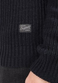 Marinblå stickad tröja med ribbat mönster och en rektangulär etikett med "JACK & Originals JONES" i vitt på en grå bakgrund.