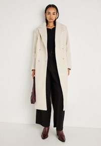 Dvojřadový kabát v krémové barvě, vyrobený z texturované tkaniny. Má široký límec, dva velké knoflíky a boční kapsy. Černé kalhoty a hnědé boty.