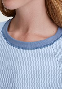 Lichtblau gestreepte stof met een gestructureerd oppervlak. De ronde halslijn heeft een effen blauwe ribbelrand. Close-up weergave van het kledingstuk.