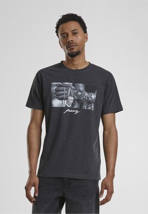 Homme portant un t-shirt gris foncé avec un graphique en noir et blanc d'une main tatouée tenant une chaîne et le mot "pray" en dessous.