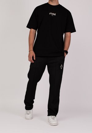Homme portant un t-shirt noir "crystal" et un pantalon noir avec un logo géométrique blanc, debout avec une main dans la poche et des baskets blanches.