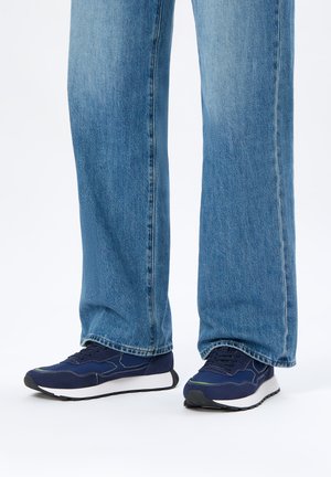 Gambe con jeans larghi blu e scarpe da ginnastica blu scuro con suola bianca su sfondo bianco.