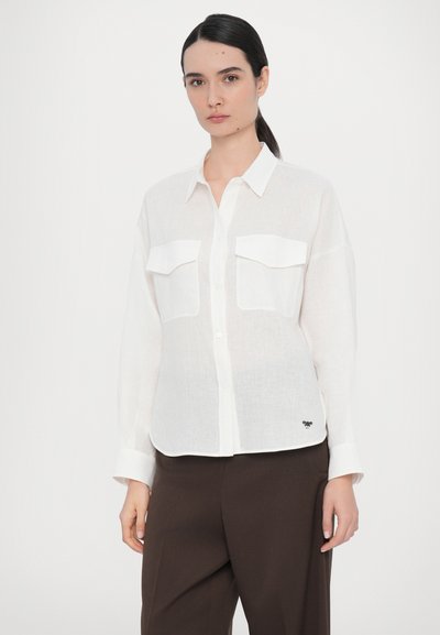 White linen button-up shirt with two chest pockets, long sleeves, and a curved hem, paired with brown trousers. Subtle branding at the hem.