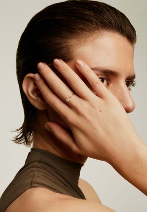 Anillo de oro con una pequeña piedra clara, llevado en la mano de un modelo; la mano está parcialmente oscurecida por los dedos y el cabello, sobre un fondo neutro.