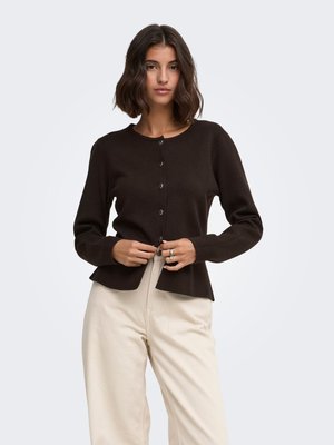 Kvinde med skulderlangt mørkt hår, der knapper en sort langærmet cardigan, iført lysebeige højtaljede bukser mod en ensfarvet baggrund.