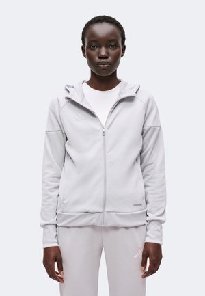 Femme portant un sweat à capuche gris clair zippé et un pantalon de survêtement assorti, debout devant un fond clair uni.