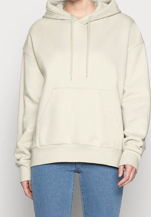 Beiger übergroßer Hoodie mit Kängurutasche vorne und verstellbarer Kapuze, getragen zu blauen Denim-Jeans, an Schultern und Hüften gekürzt.