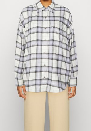 Personne portant une chemise boutonnée oversized avec un motif à carreaux blanc, noir et lavande, associée à un pantalon beige.