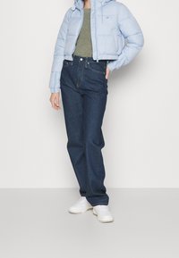 Osoba oblečená v světle modré kratké péřové bundě, zeleném pleteném topu, tmavě modrých džínách s vysokým pasem a bílých teniskách stojící na bílém pozadí.