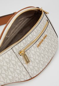 Sac bandoulière en tissu crème avec un motif géométrique. Accents en cuir marron, quincaillerie dorée et compartiments zippés. Détail de logo remarquable.