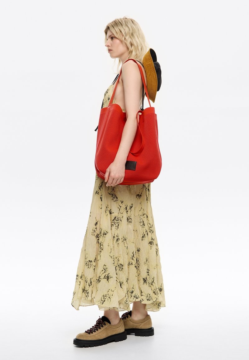 Rode tote bag met een gestructureerd oppervlak, gecombineerd met een gele bloemenjurk en beige schoenen. De tas heeft dubbele handvatten en een logodetail.