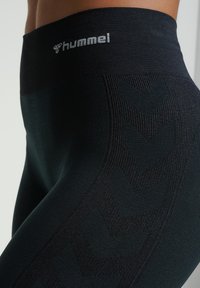 Zbliżenie osoby noszącej czarne legginsy marki Hummel z teksturowanym wzorem w jodełkę po bokach i szerokim elastycznym paskiem w talii.