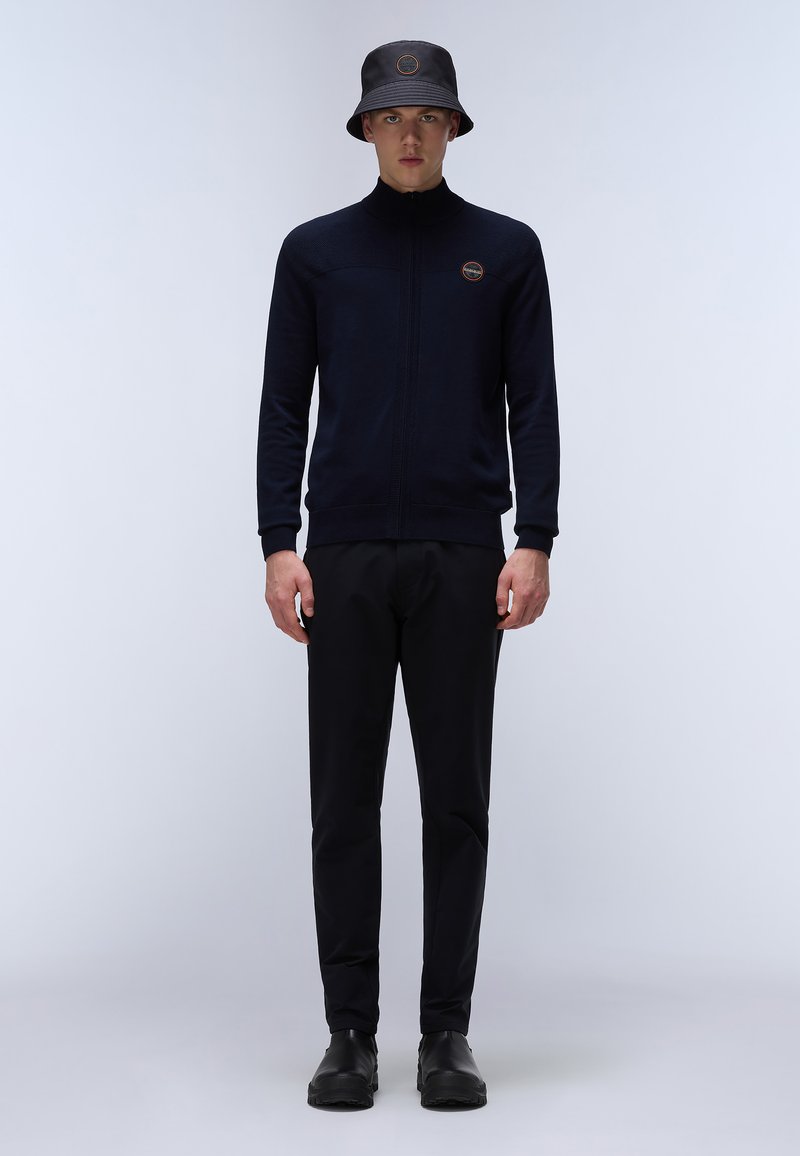 Sweat à zip bleu marine avec un design texturé, associé à un pantalon noir et un chapeau seau noir arborant un logo. Couleurs unies, coupe décontractée.