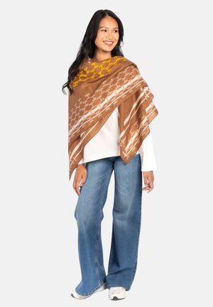 Vrouw met een bruin patroon sjaal over een wit shirt met lange mouwen, blauwe wijde jeans en witte sneakers, glimlachend en staand tegen een witte achtergrond.