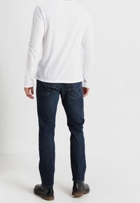 Camisa blanca de manga larga y vaqueros azul oscuro combinados con botas negras, vistos desde atrás, mostrando el diseño sencillo de la camisa y el corte clásico de los vaqueros.