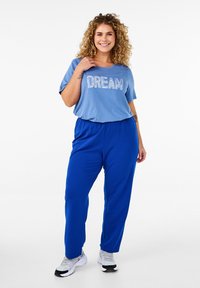 Světle modré tričko s textem "DREAM", spárované s královsky modrými kalhotami s elastickým pasem. Model má na sobě bílé tenisky a stojí na neutrálním pozadí.