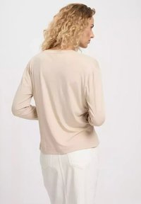 Beżowa bluzka z długim rękawem o luźnym kroju. Wykonana z miękkiego materiału, z okrągłym dekoltem i prostym wzornictwem. Noszona z białymi spodniami, widoczna kratkowana struktura.