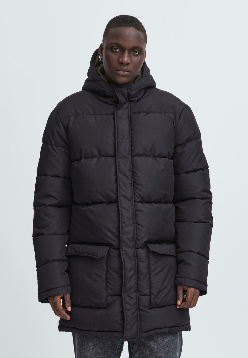 Abrigo acolchado negro con capucha, que presenta un diseño acolchado, dos bolsillos frontales y un zipper de longitud completa. La tela tiene una textura suave.