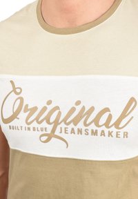 Bež i boja slonovače majica s okruglim izrezom, s natpisom "Original Built in Blue Jeansmaker" u svijetlosmeđem fontu na prsima.