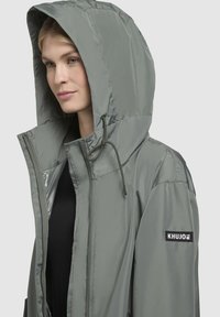 Jachetă cu glugă de culoare gri deschis, cu fermoar pe față, glugă cu șnur și patch cu logo pe mâneca stângă. Textură netedă și strălucitoare, cu un design aerodinamic.
