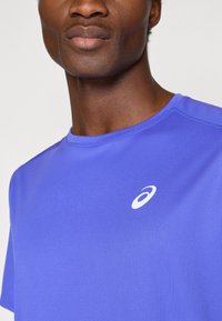 Μωβ αθλητικό μπλουζάκι με κοντά μανίκια, φτιαγμένο από ελαφρύ ύφασμα. Διαθέτει λευκό λογότυπο στο στήθος και στρογγυλή λαιμόκοψη.