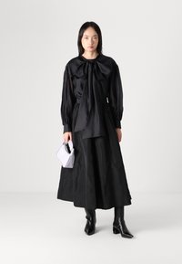 Zwarte lange mouwen jurk met strikdetail bij de nek, uitlopende rok, gecombineerd met een kleine witte handtas en zwarte puntige laarzen.