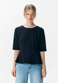 Zwarte top met korte mouwen en een ronde halslijn, gemaakt van een soepele stof. Heeft een relaxed fit en subtiele plooien bij de taille. Gekleed met lichtblauwe jeans.