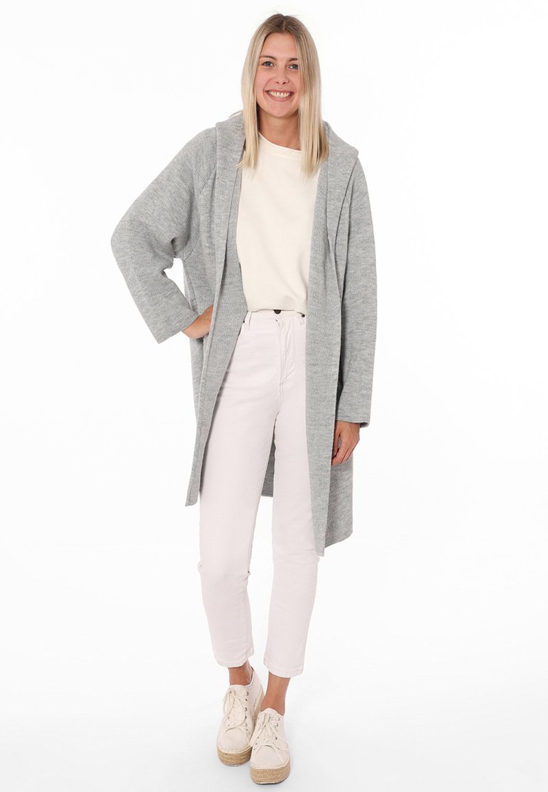 Cappotto grigio a maniche lunghe, con cappuccio e vestibilità ampia, indossato sopra una maglietta bianca e pantaloni rosa chiaro, abbinato a espadrillas beige.