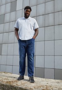 Polo à manches courtes gris clair avec un logo rouge, associé à un jean en denim bleu et des chaussures noires à lacets, devant un mur carrelé gris.