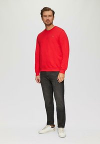 Sweat-shirt rouge avec encolure ras du cou, poignets côtelés et ourlet. Porté avec un jean foncé et des baskets blanches avec des accents noirs. Design simple.
