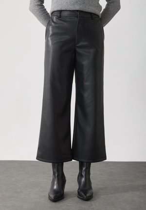 Personne portant un pantalon en cuir noir à coupe large associé à des bottines noires et un pull gris, debout sur un sol gris devant un fond blanc.