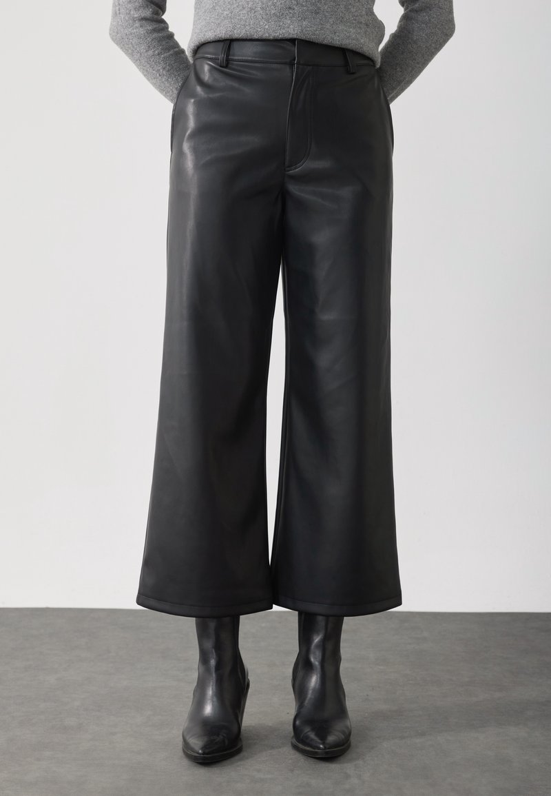 Personne portant un pantalon en cuir noir à coupe large associé à des bottines noires et un pull gris, debout sur un sol gris devant un fond blanc.
