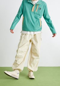 Pullover in pile di un tenue blu verde con colletto a bottoni e dettagli beige, abbinato a pantaloni cargo bianco sporco e scarpe alte beige.