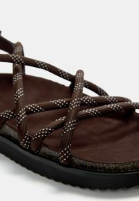 Barna szandál keresztbe font textile pántokkal, fehér akcentusokkal, puha barna talppal és texturált fekete gumitalppal.
