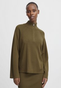 Donna con capelli corti che indossa un maglione zip-up verde oliva e una gonna a coste abbinata, in piedi davanti a uno sfondo bianco semplice.