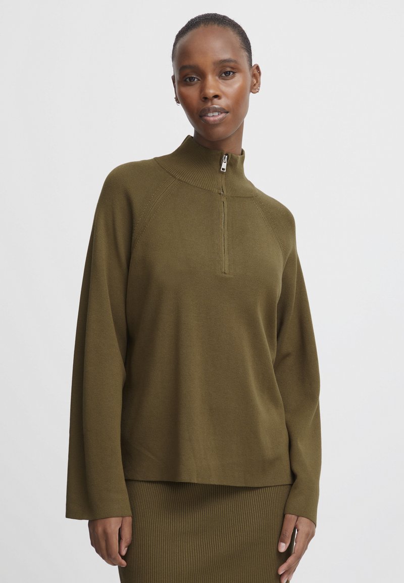 Donna con capelli corti che indossa un maglione zip-up verde oliva e una gonna a coste abbinata, in piedi davanti a uno sfondo bianco semplice.