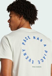 Lys grøn t-shirt med korte ærmer, der har blå cirkulær tekst "EVERY PACE HAS A PLACE" og "ADIDAS RUNNERS" i grøn nederst.