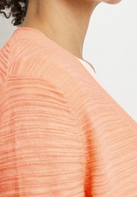 De nek en schouder van een persoon die een textuur, perzikkleurig gebreid kledingstuk draagt, tegen een effen witte achtergrond.