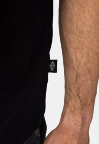 Černé bavlněné tričko s bočním štítkem, na kterém je bílá geometrická značka. Materiál vypadá měkce a má hladkou texturu.