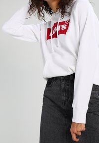 Sudadera blanca con un logo rojo de Levi's en el frente, que cuenta con un bolsillo canguro y capucha con cordón, combinada con una falda de mezclilla negra.